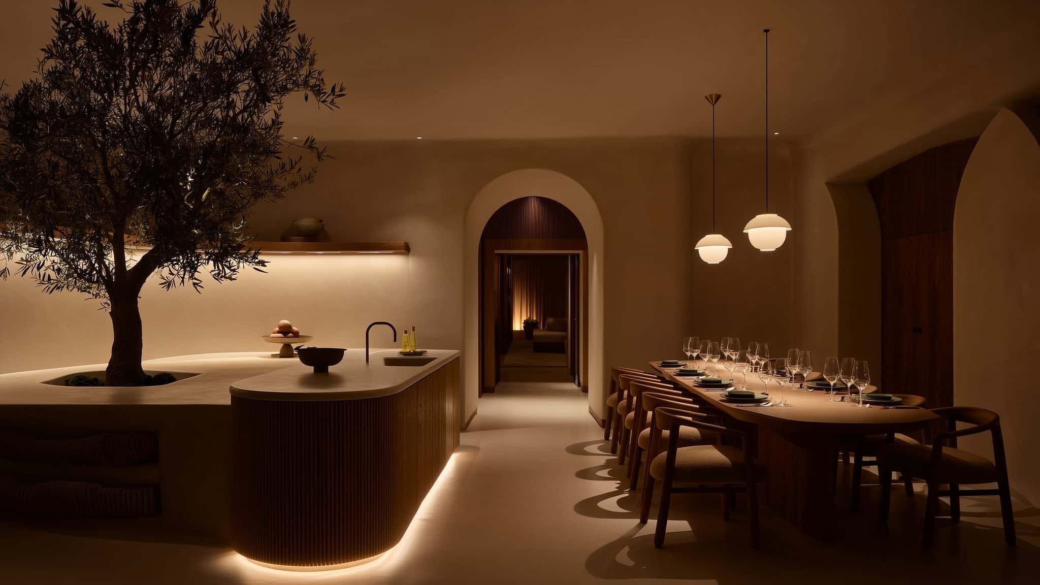 Warm plaster dining room at dusk with an arched passage, an olive tree, and low pooled light